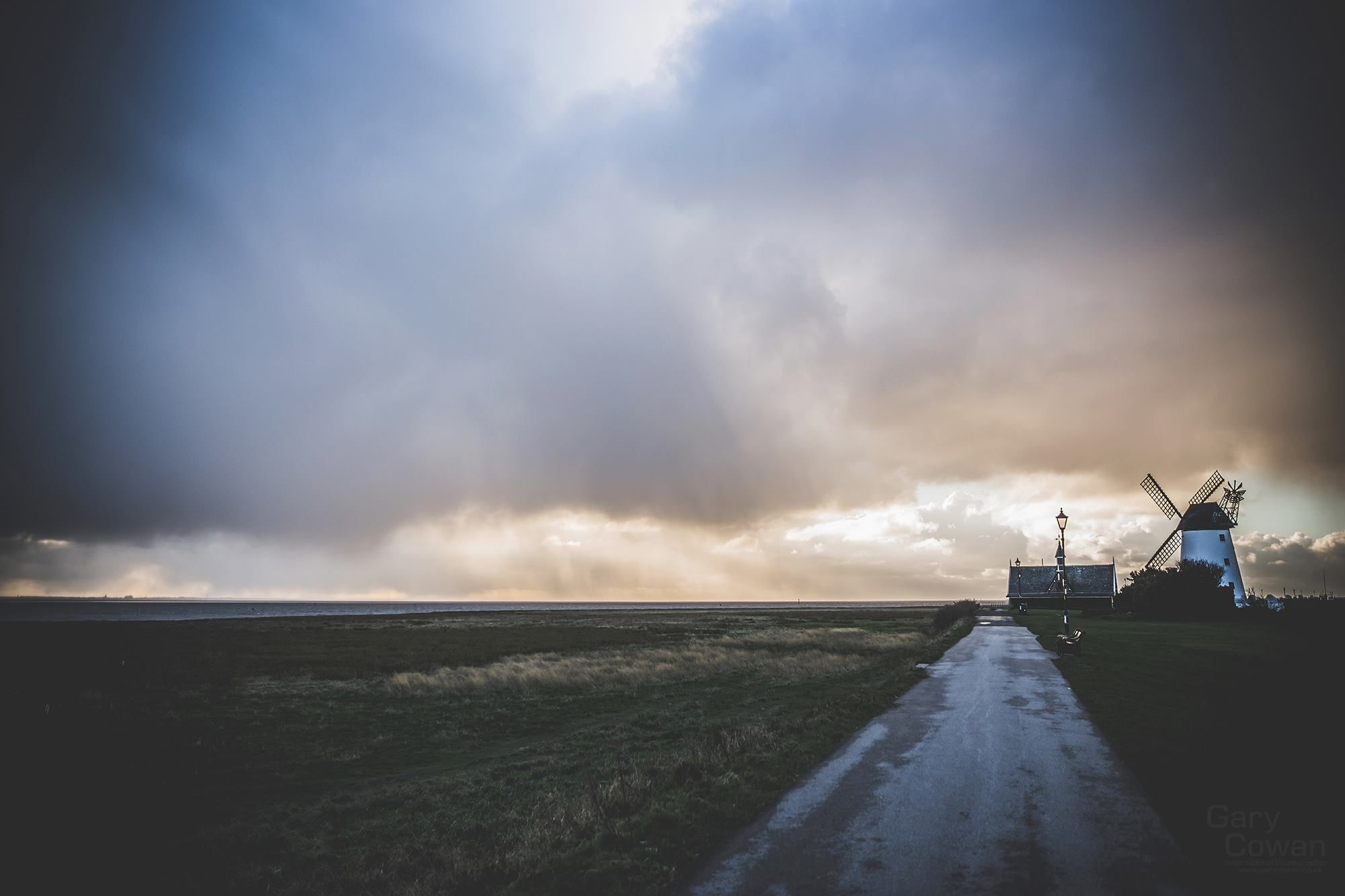 Sunlight breaking through dark clouds above a coastal path leading towards a white windmill