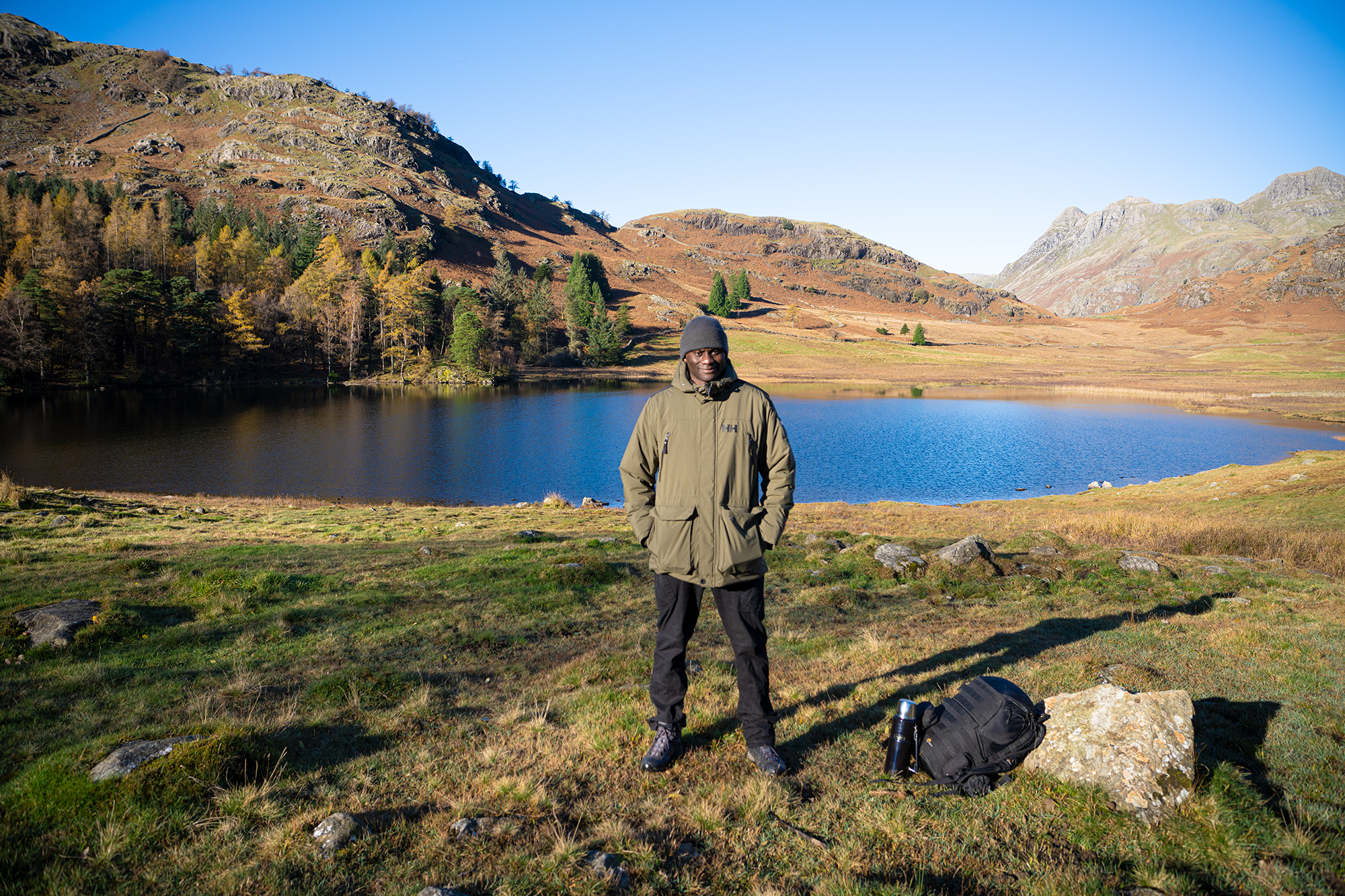 A walker standing beside a lake beneath rocky hills, with a backpack and flask on the grass