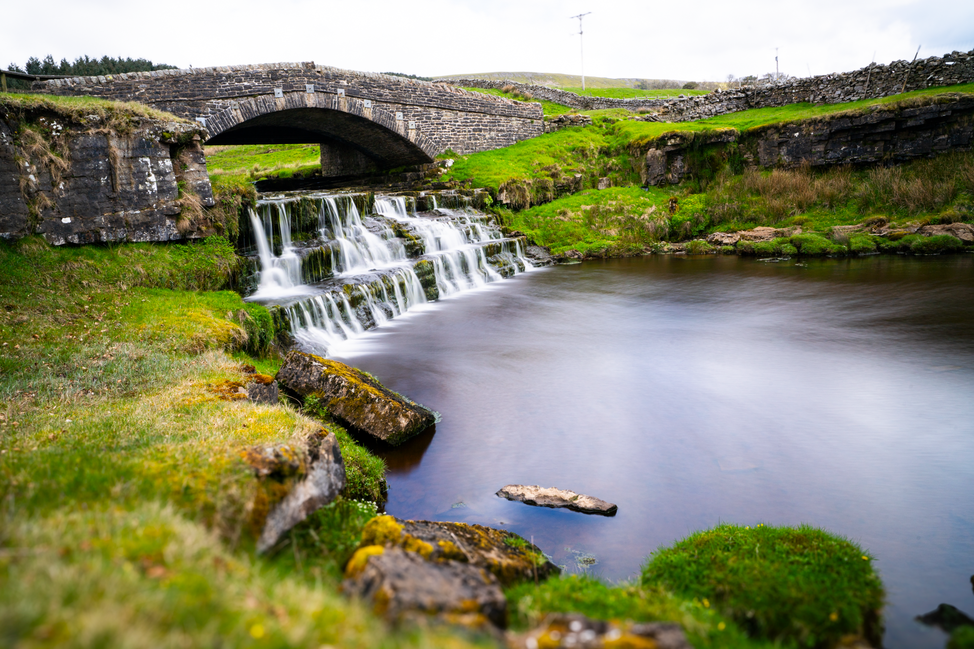 A stone arch bridge above a stepped waterfall and a calm pool, surrounded by green grass