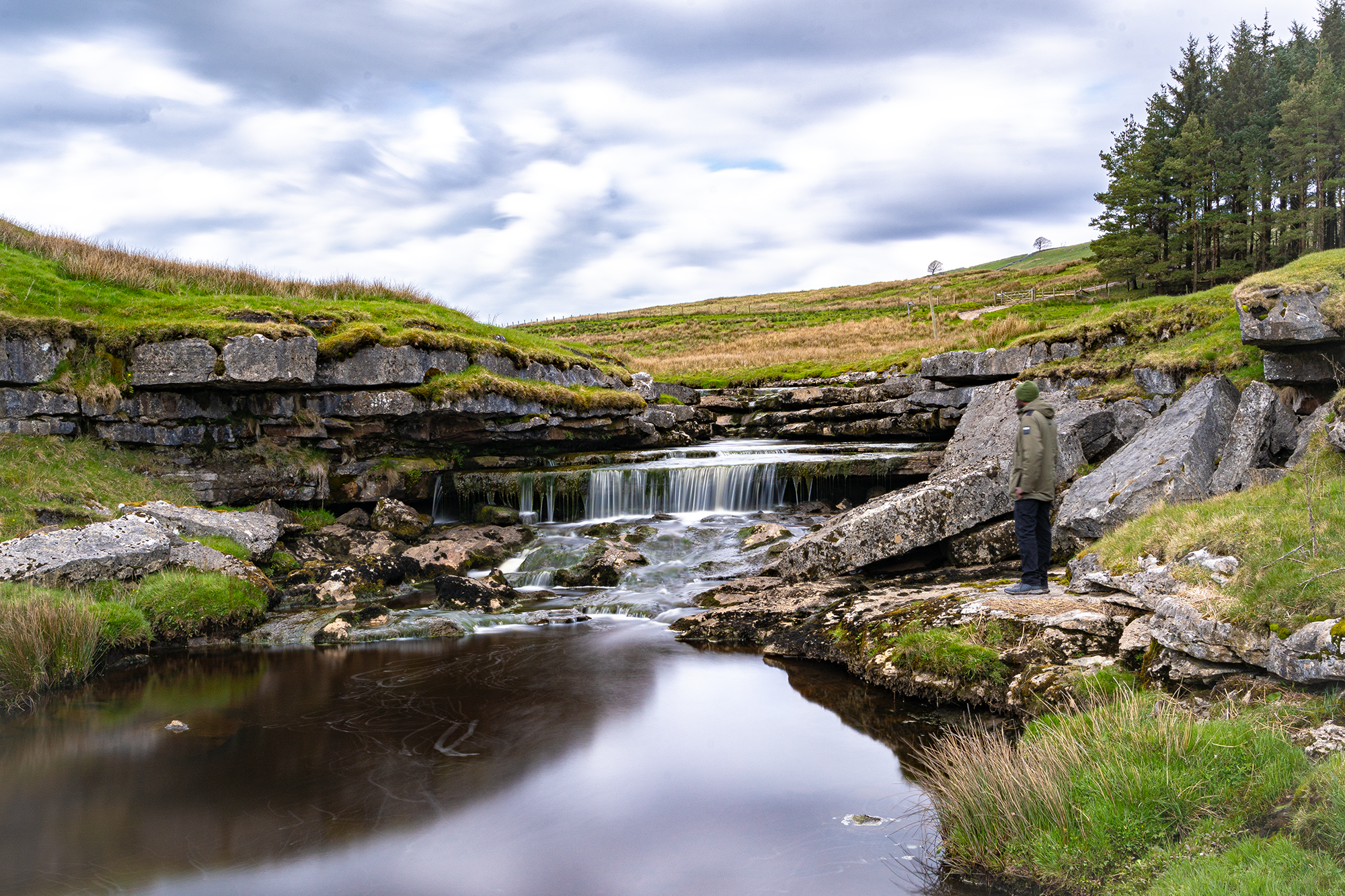 A person standing on rocks beside a stepped waterfall below grassy slopes and trees