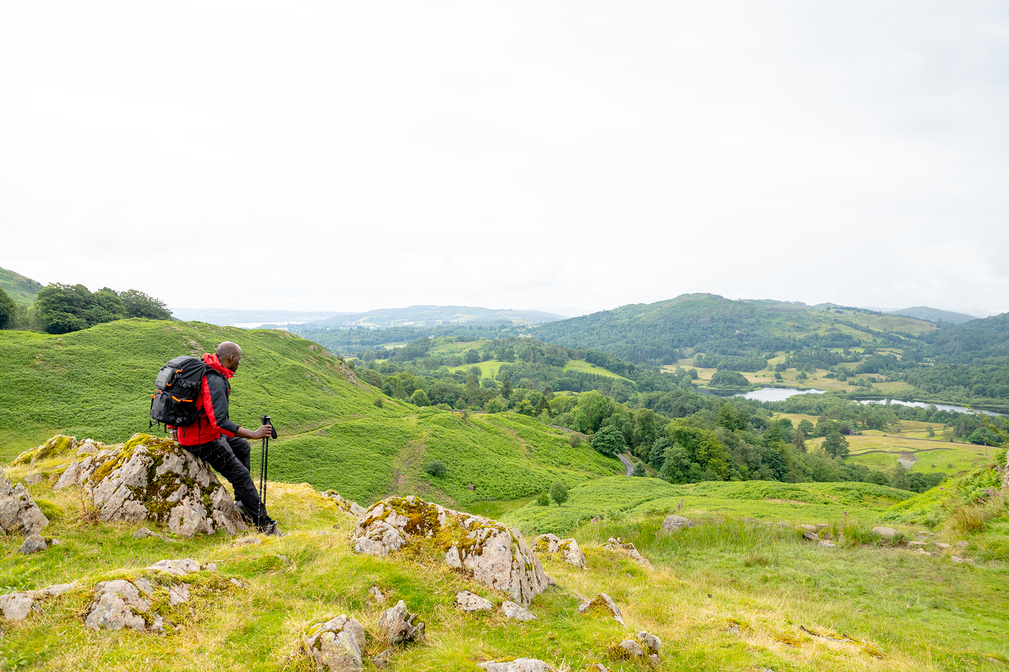 A walker with a backpack and walking poles resting on a rocky hillside above green fields, trees and water