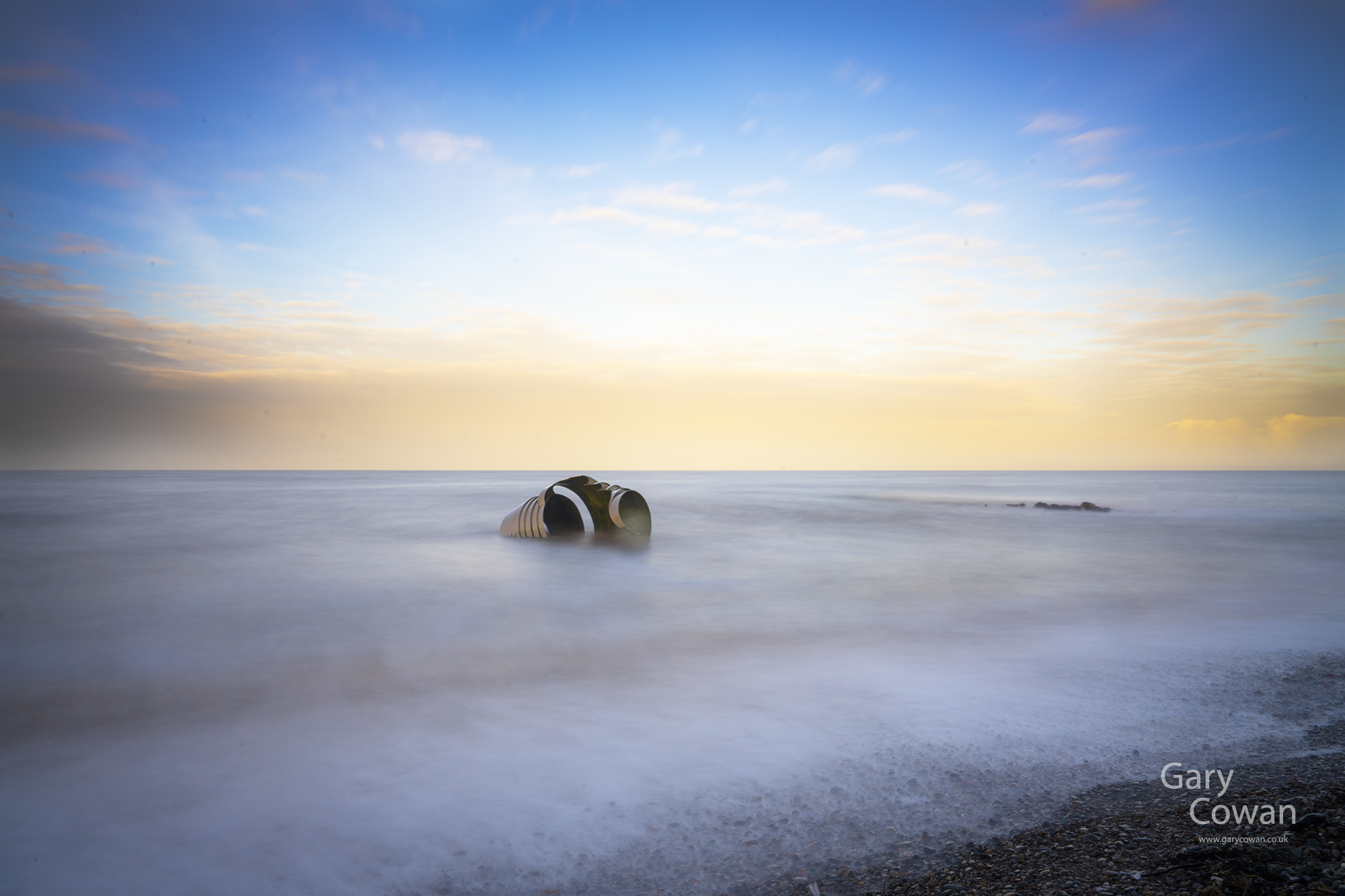A curved metal sculpture surrounded by softly blurred seawater beneath a pale blue and golden sky